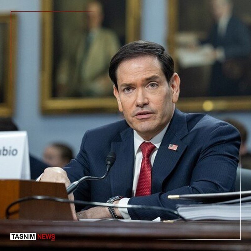 A dark-haired man in a navy suit and red tie sits at a wooden desk with a microphone, wearing an American flag lapel pin, with framed portraits visible in the background.