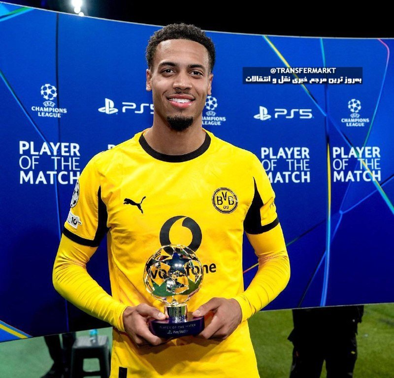 A smiling soccer player in a yellow Borussia Dortmund jersey holds a Champions League Player of the Match trophy in front of a branded backdrop.