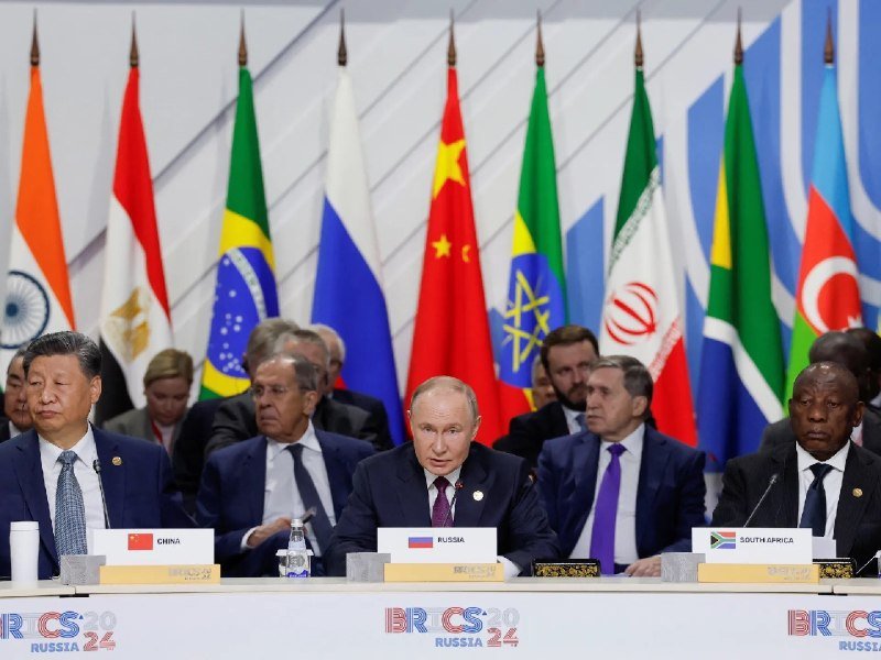 Several men in suits sit at a labeled table before a row of national flags, with a "BRICS RUSSIA 2024" banner displayed along the front.