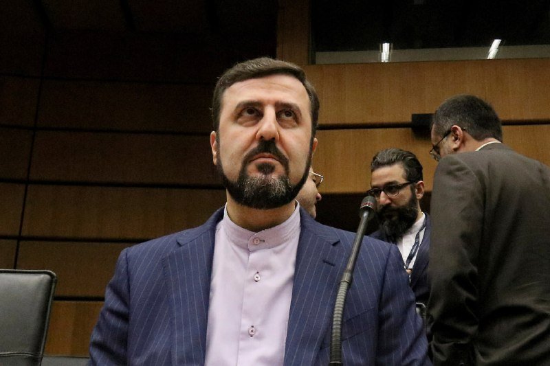A bearded man in a blue pinstripe suit and lavender shirt stands near a microphone in a wood-paneled room, with two other men visible behind him.