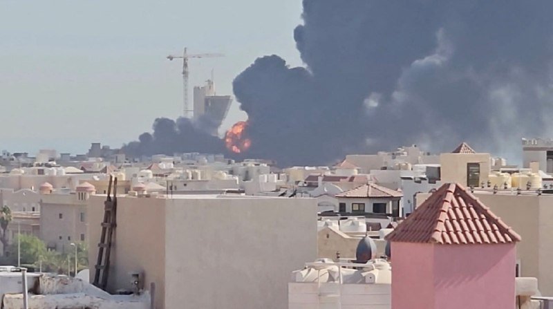 A large plume of thick black smoke and visible orange flames rise above a densely packed cityscape of low-rise buildings, with a construction crane visible in the background.