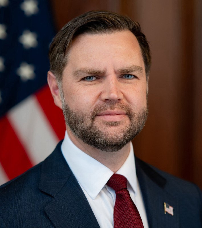 A bearded man in a dark suit, white shirt, and red tie poses for an official portrait with an American flag and a wooden backdrop behind him.