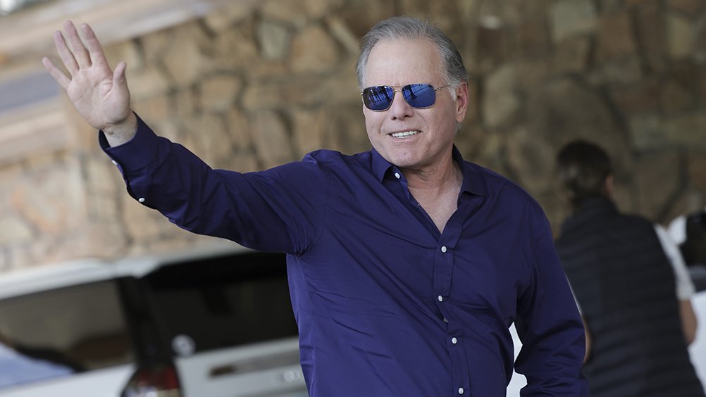 A gray-haired man wearing blue sunglasses and a dark blue button-up shirt smiles and waves toward the camera, standing outdoors near a white vehicle and another person.