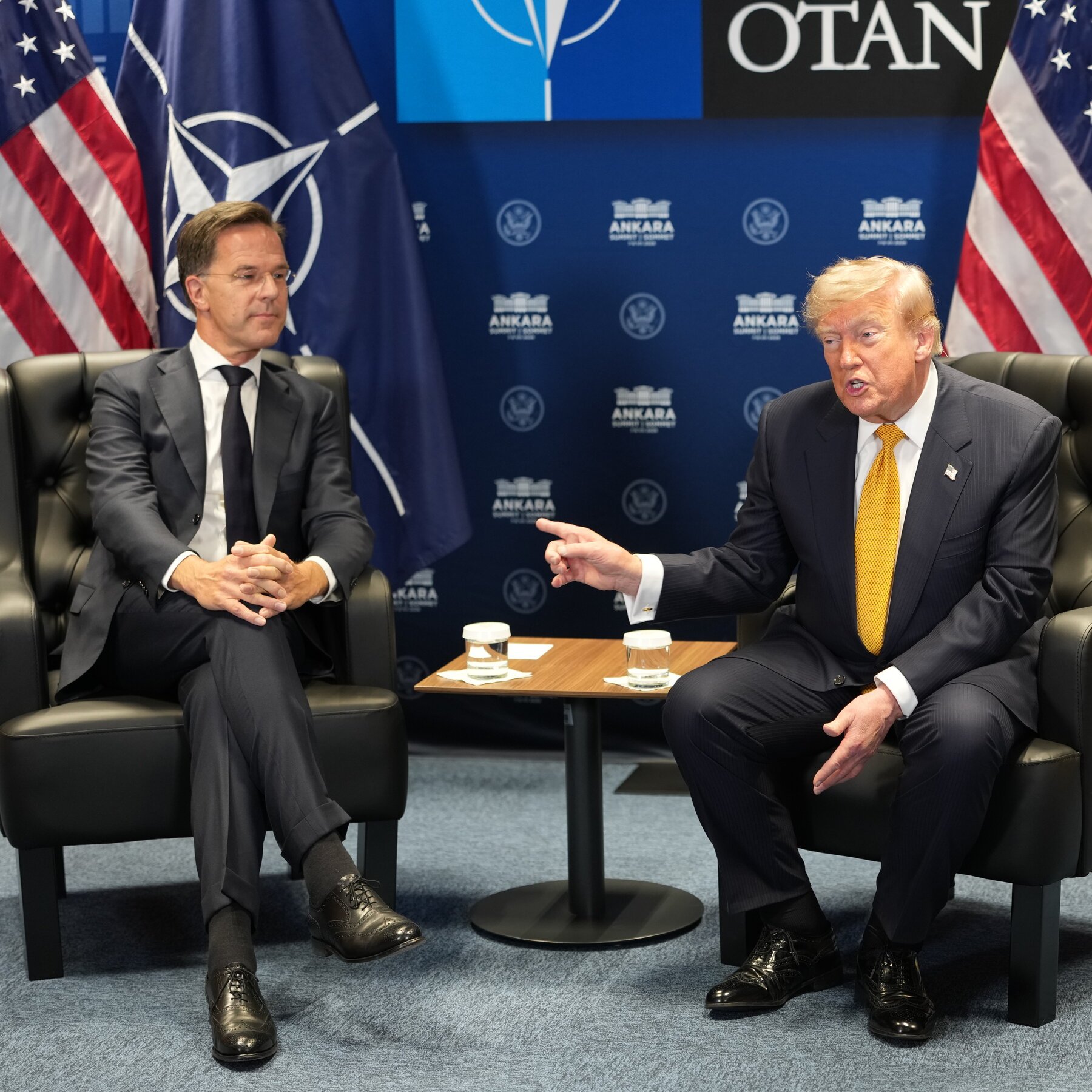 Two men in suits sit in black armchairs facing each other, with one gesturing while speaking, in front of a blue backdrop displaying NATO and Ankara Summit logos flanked by American flags.