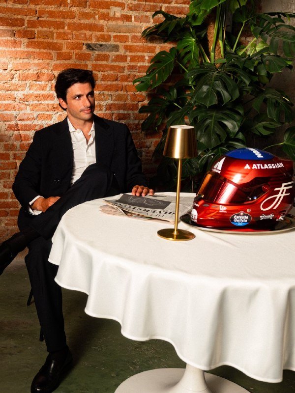A man in a dark suit sits at a white-clothed round table beside a red Atlassian-branded racing helmet, a gold lamp, and a large green plant against an exposed brick wall.