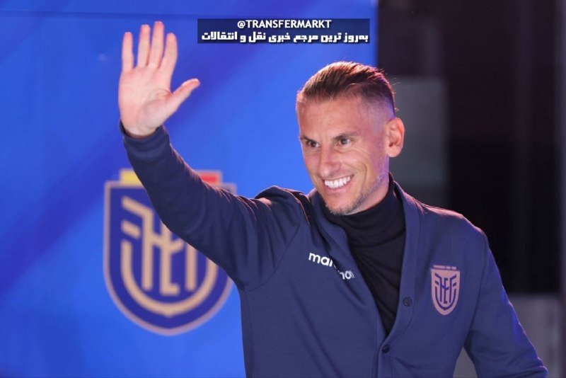 A smiling man in a navy blazer with a Macron logo waves at the camera in front of a blue wall displaying a shield-shaped crest and Transfermarkt watermark text.