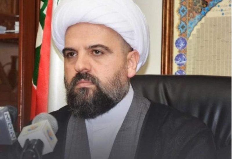 A bearded man in a white turban and black robes sits before microphones, with a flag visible in the background.