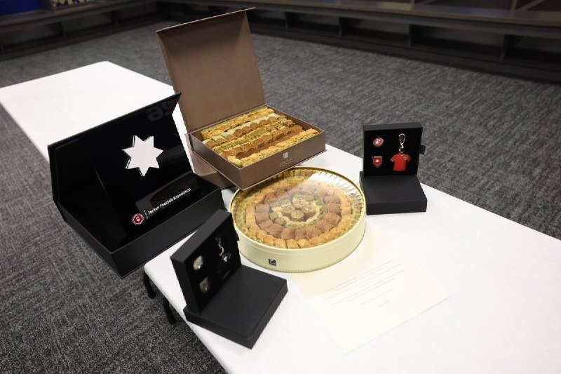 A white table displays boxed pastries, a round container of assorted baklava, a keychain with a red jersey, and a printed document in an indoor setting.