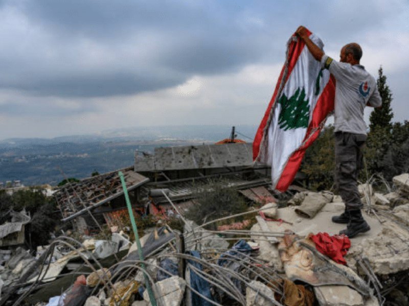 A man waves a Lebanese flag amid extensive rubble from collapsed buildings on a hilltop under a cloudy sky.