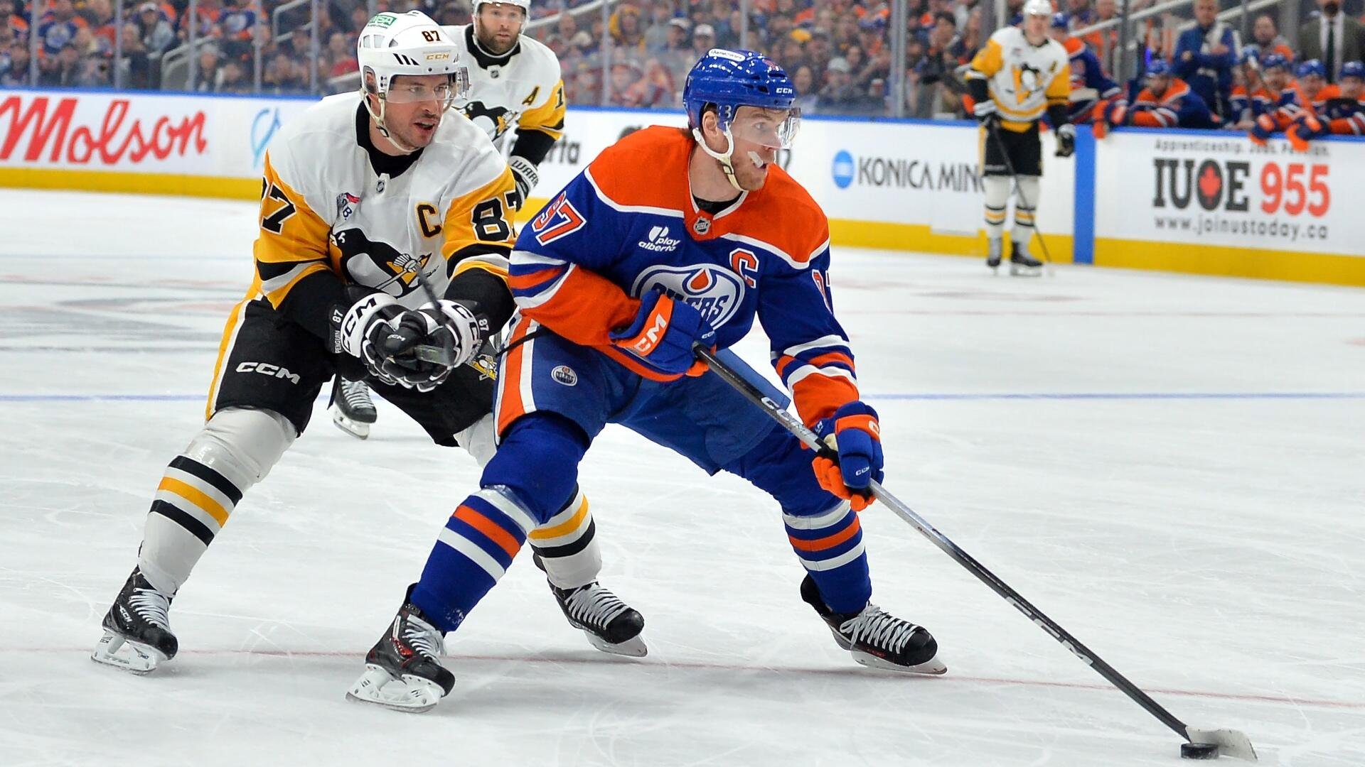 A hockey player in a blue and orange jersey skates with the puck while being defended by an opponent in a white and yellow jersey during a game.