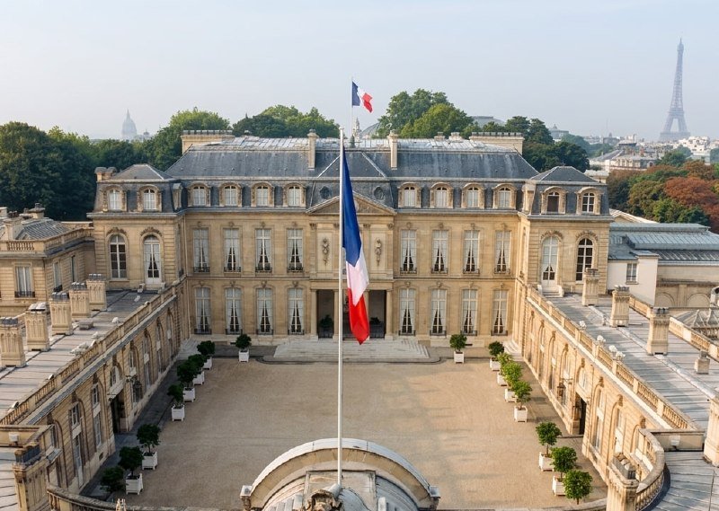 A French flag flies at half-mast on a tall pole in the courtyard of a grand classical stone building, with the Eiffel Tower visible in the background.