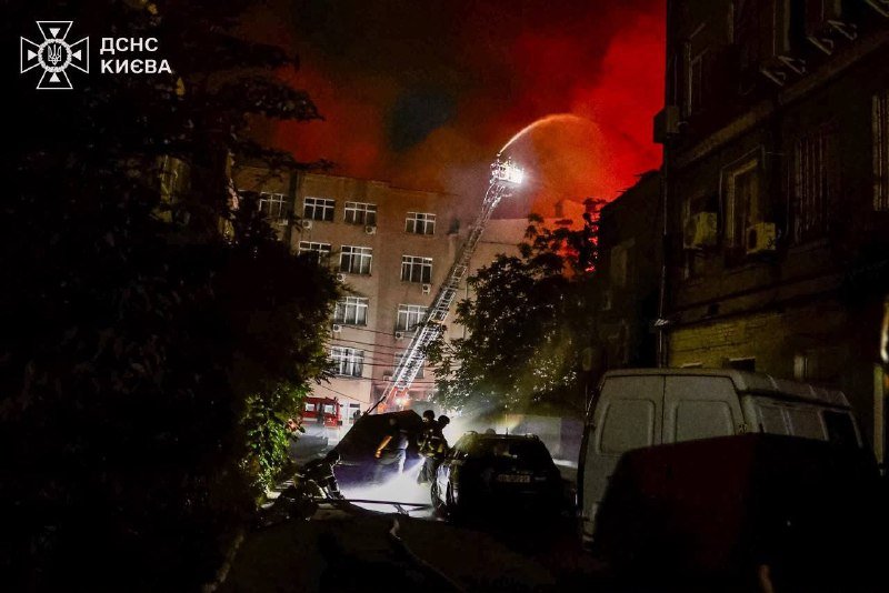 Firefighters use an extended aerial ladder to spray water onto a burning multi-story building at night, with smoke glowing red and emergency vehicles parked below.