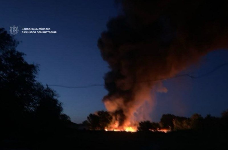 A large fire burns at ground level at dusk, producing a tall plume of dark smoke rising into the sky, with trees silhouetted in the foreground.