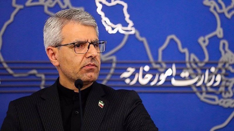 A gray-haired man wearing glasses and a dark suit speaks into a microphone in front of a blue backdrop featuring a map outline and Persian script.