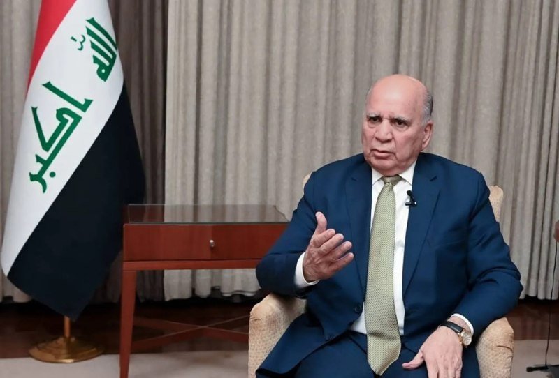 A bald man in a blue suit and green tie gestures with his right hand while seated, with an Iraqi flag visible to his left against beige curtains.
