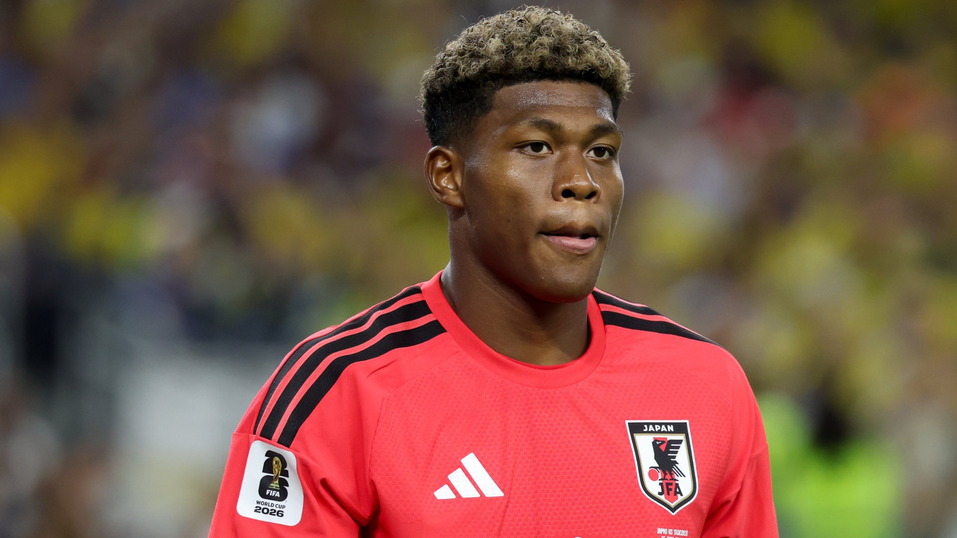 A young soccer player with bleached curly hair stands on the field wearing a red Japan national team jersey featuring the JFA crest and a FIFA World Cup 2026 qualifier patch.