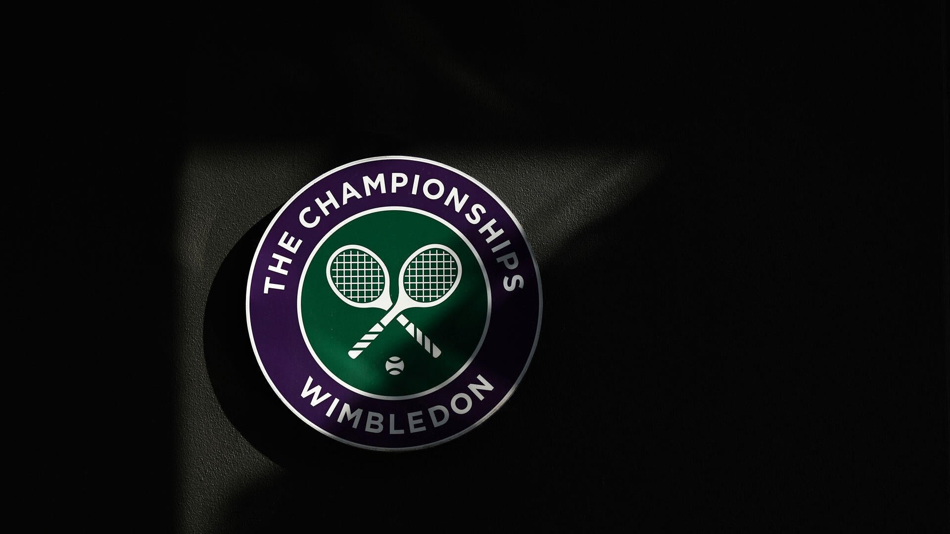 The circular Wimbledon Championships logo, featuring crossed tennis rackets and a ball on a purple and green background, is displayed against a dark backdrop.