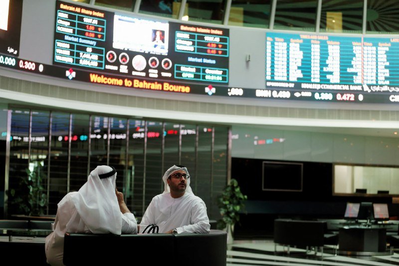 Two men in traditional white Arab attire converse while seated at a desk beneath large digital displays reading "Welcome to Bahrain Bourse" and showing stock market data.