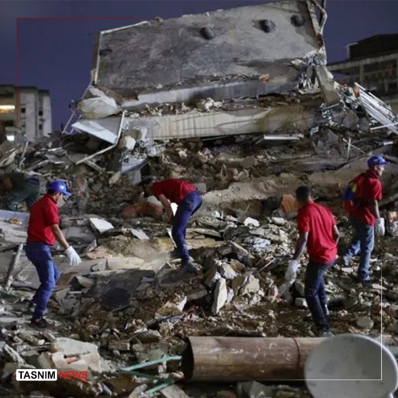 Rescue workers in red shirts and blue caps search through the rubble of a collapsed concrete building at night.