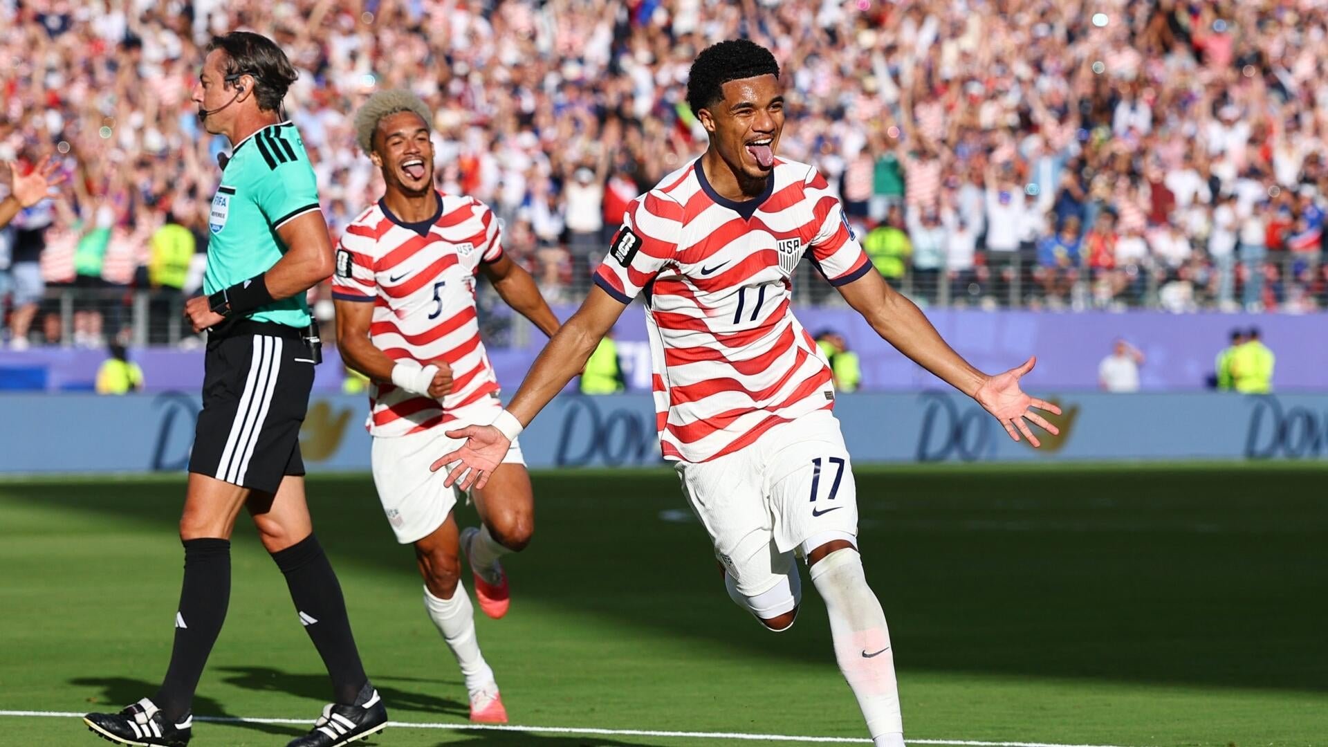 Soccer players in red-and-white striped USA jerseys, including numbers 3 and 17, celebrate on the pitch near a referee while a large crowd cheers from the stands.