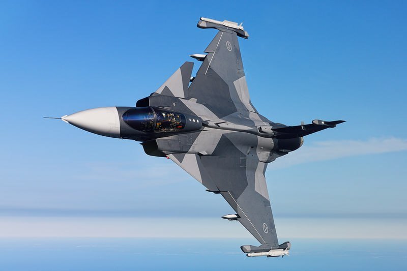 A gray fighter jet with wingtip missiles banks sharply against a clear blue sky, its cockpit canopy reflecting sunlight.