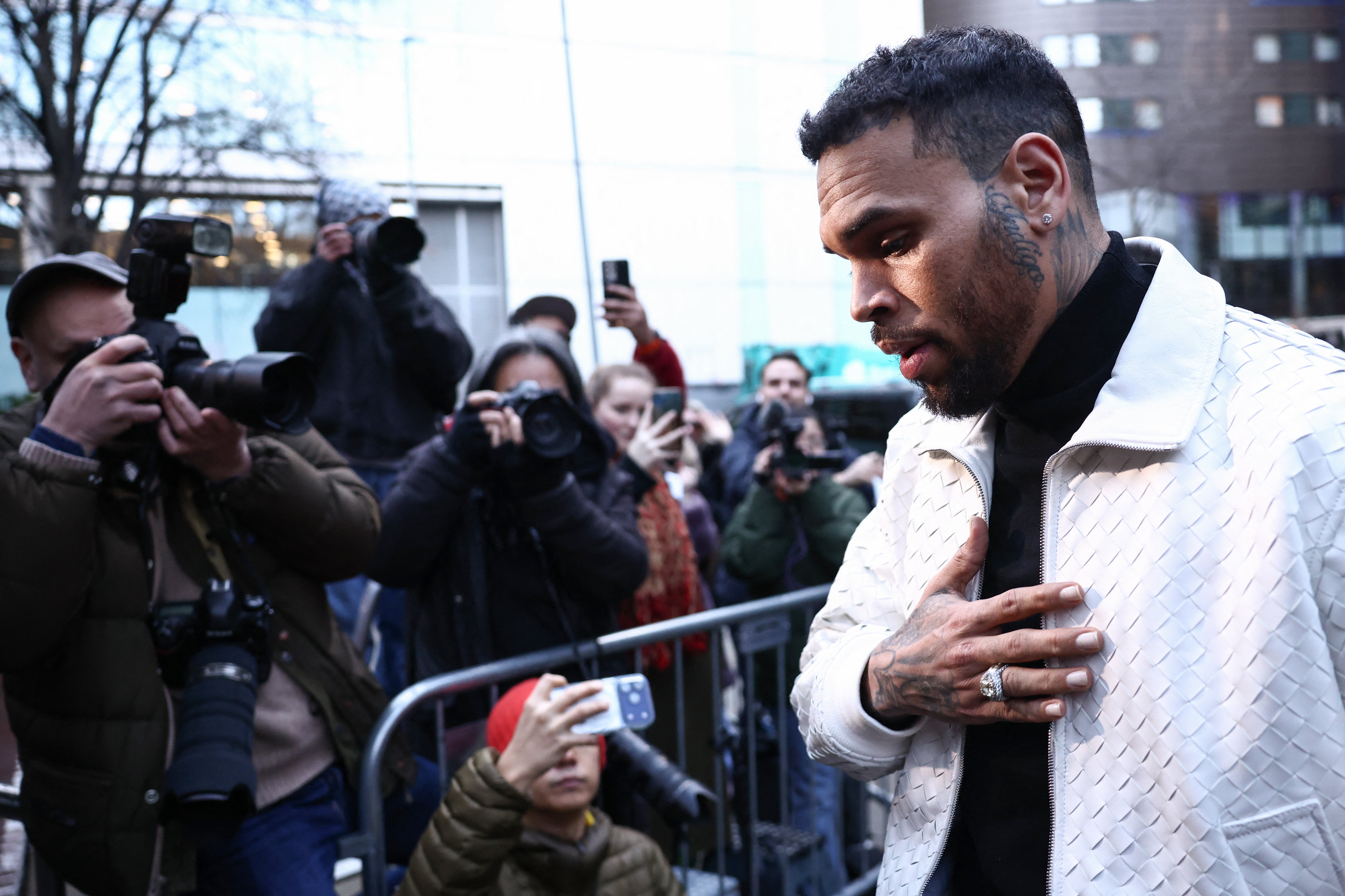 A bearded man with face tattoos wearing a white quilted jacket places his hand on his chest while photographers take pictures from behind a metal barricade.