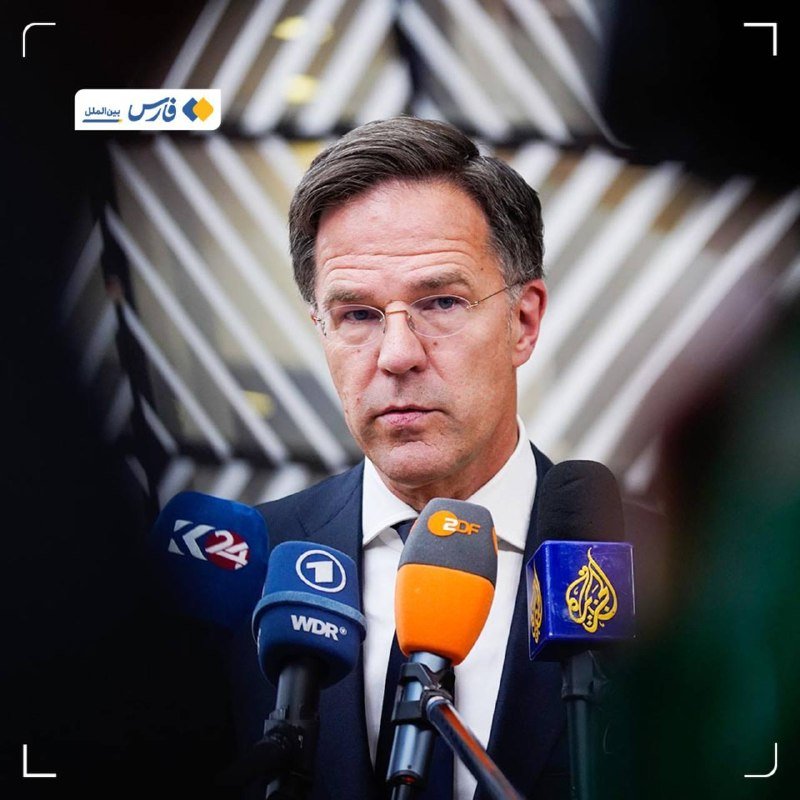 A man in a suit and glasses speaks into multiple microphones labeled "24," "WDR," "ZDF," and Al Jazeera, with a striped backdrop behind him.
