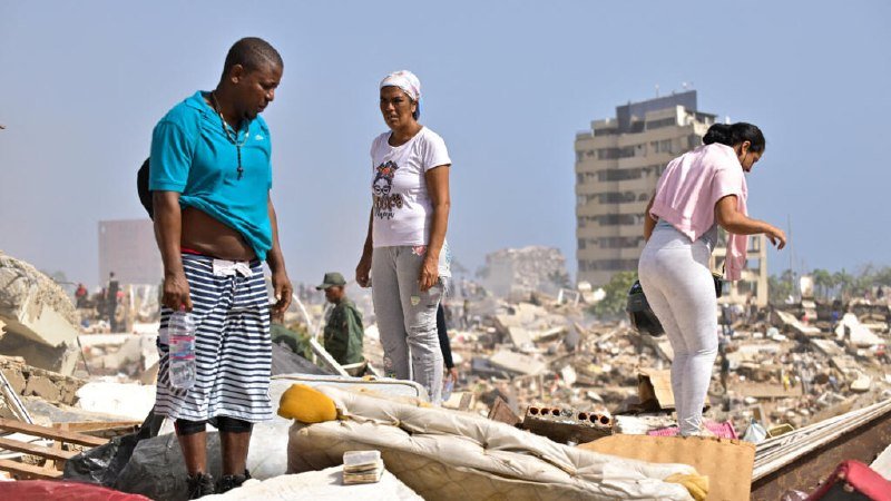 Three people stand amid scattered debris and ruined furniture, with a damaged multi-story building visible in the hazy background.