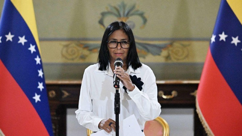 A woman with dark hair and glasses speaks into a microphone, flanked by two Venezuelan flags in front of an ornate wall.
