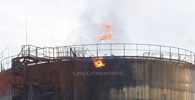 Flames and thick smoke rise from a rusted industrial storage tank surrounded by a metal railing.
