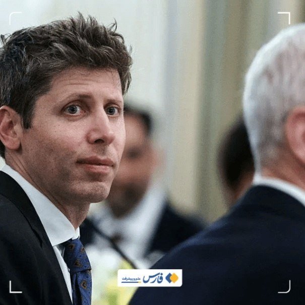 A dark-haired man in a black suit and patterned tie looks toward another man in a dark suit, with additional people visible in the background.