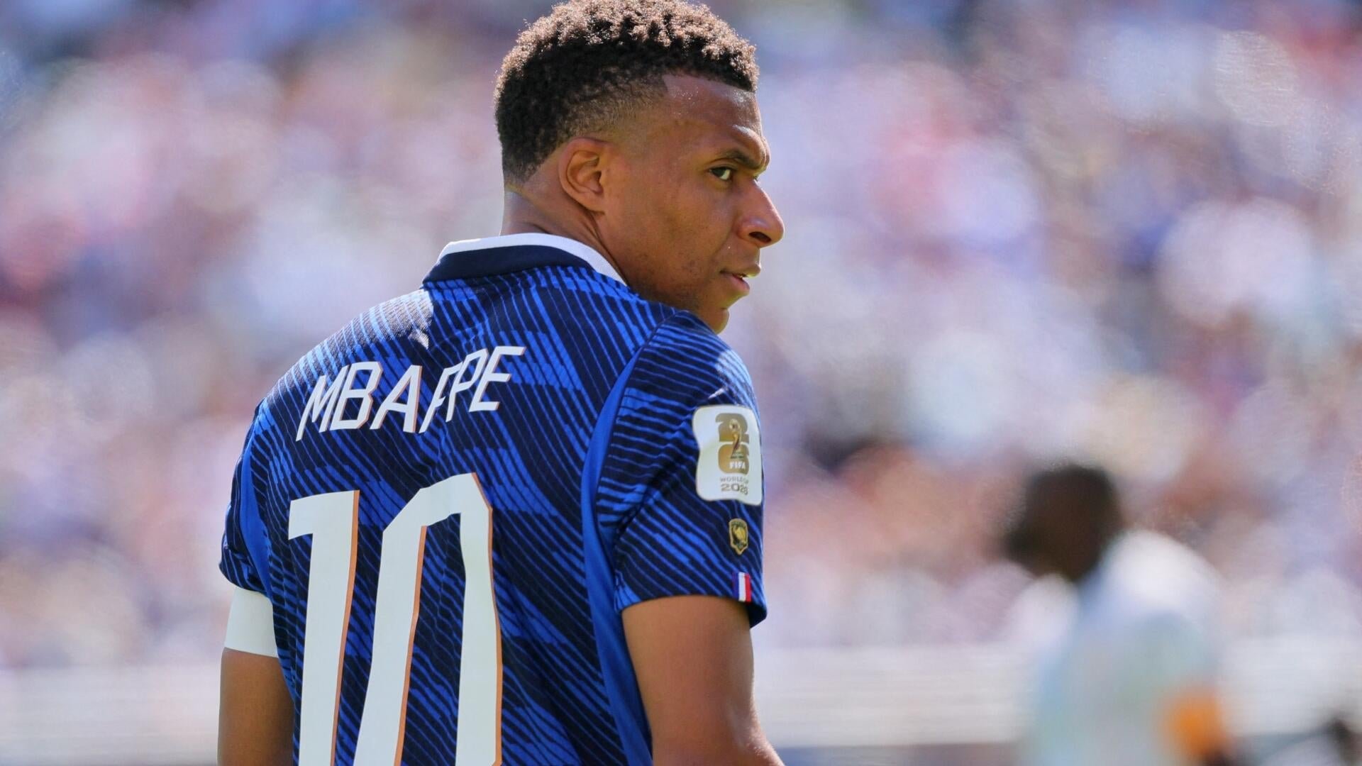 A soccer player wearing a blue jersey with "MBAPPE" and the number 10 on the back looks over his shoulder in a stadium filled with spectators.