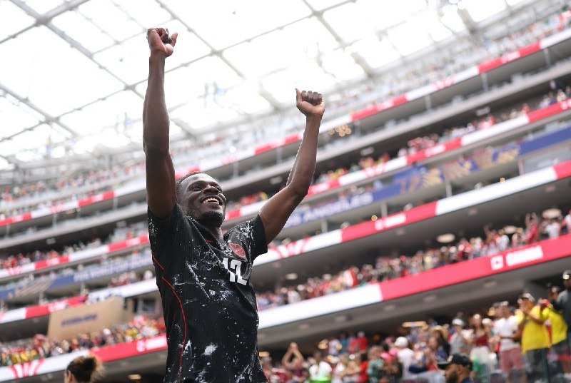 A smiling soccer player wearing a black jersey raises both arms triumphantly inside a packed stadium.