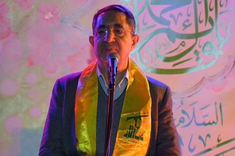 A bespectacled man wearing a yellow sash stands at a microphone, smiling in front of a backdrop featuring Arabic calligraphy and pink floral designs.