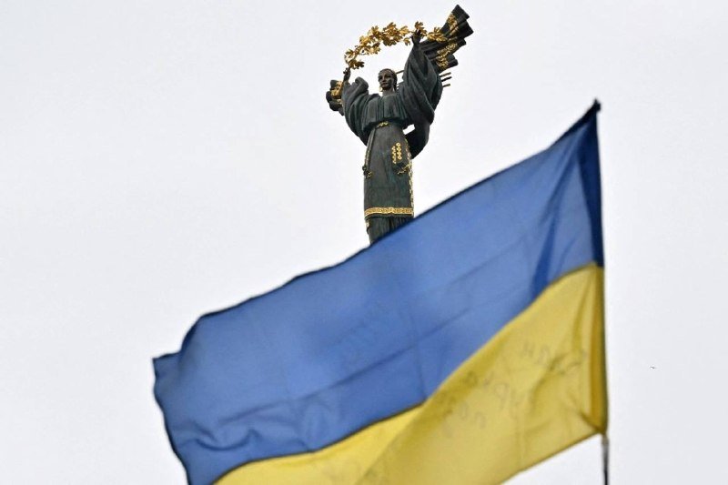 A bronze statue holding a golden wreath stands above a blue and yellow Ukrainian flag waving against an overcast sky.