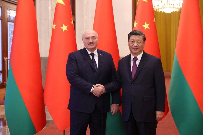 Two men in dark suits shake hands in front of Chinese and Belarusian flags inside an ornate room.