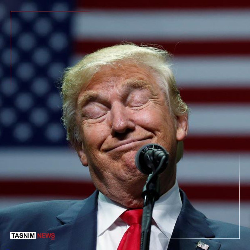 A man with blonde hair in a dark suit and red tie stands at a microphone with his eyes closed, an American flag blurred in the background.