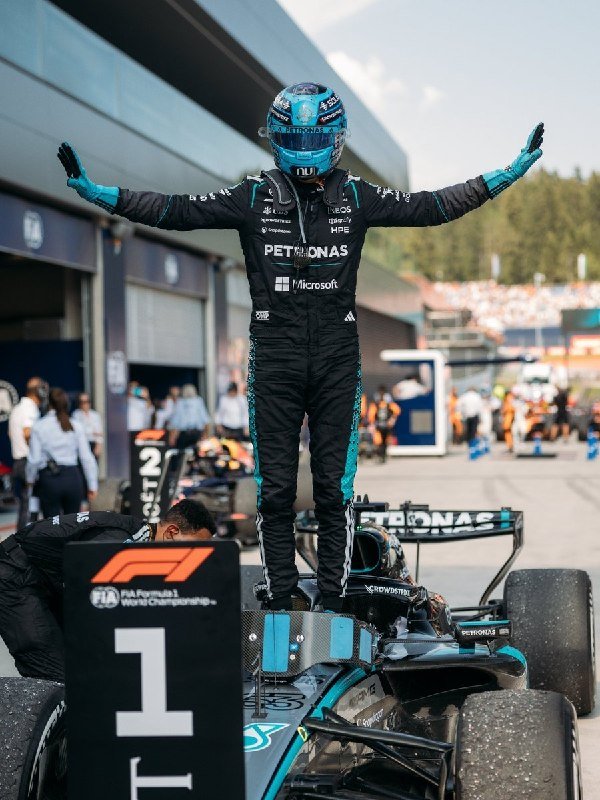 A race car driver in a black and teal Petronas racing suit stands atop a black and teal Formula 1 car with arms raised in victory, marked "1st Place" near a pit lane.
