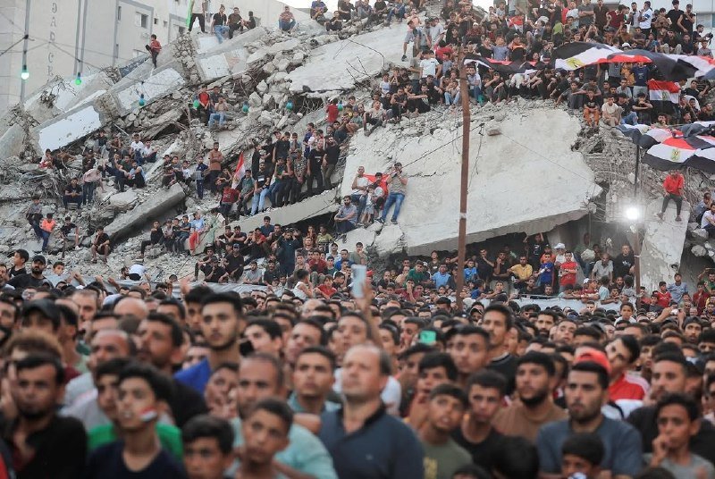 A large crowd of people gathers around and atop a massive pile of concrete rubble from a destroyed building, with some individuals holding flags and umbrellas.