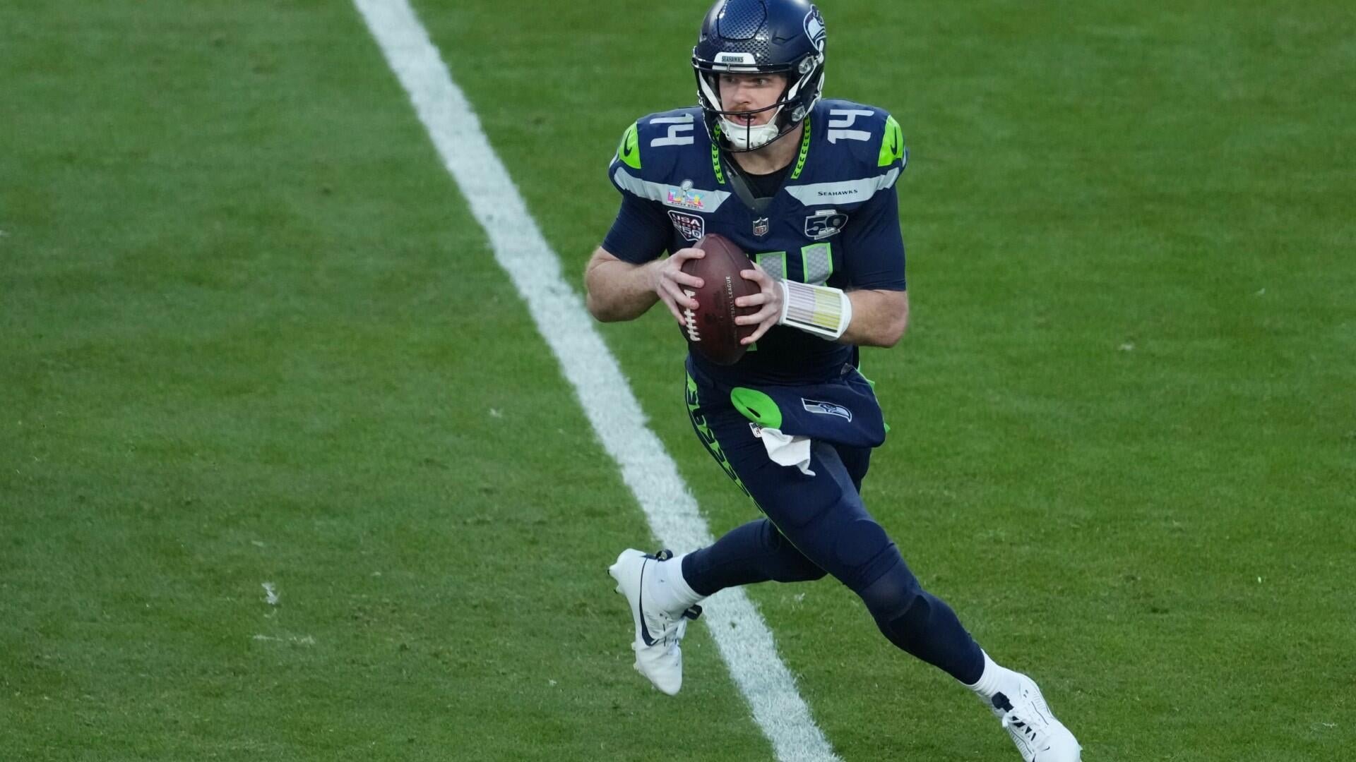 A football player wearing a navy blue and green Seahawks uniform, number 12, runs across a grass field holding a football.