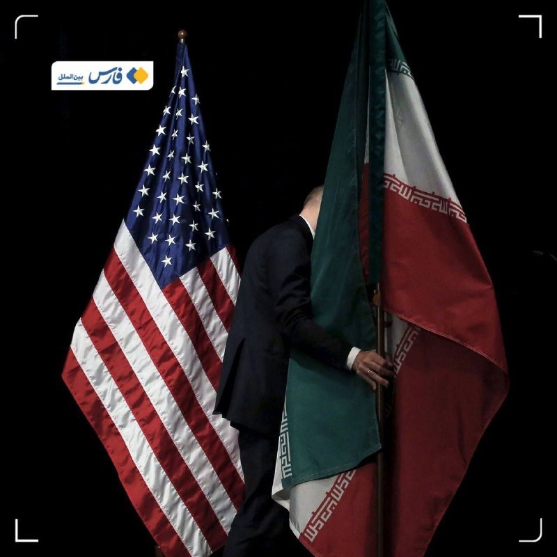 A man in a dark suit stands between the American flag and the Iranian flag, with his head bowed behind the Iranian flag.