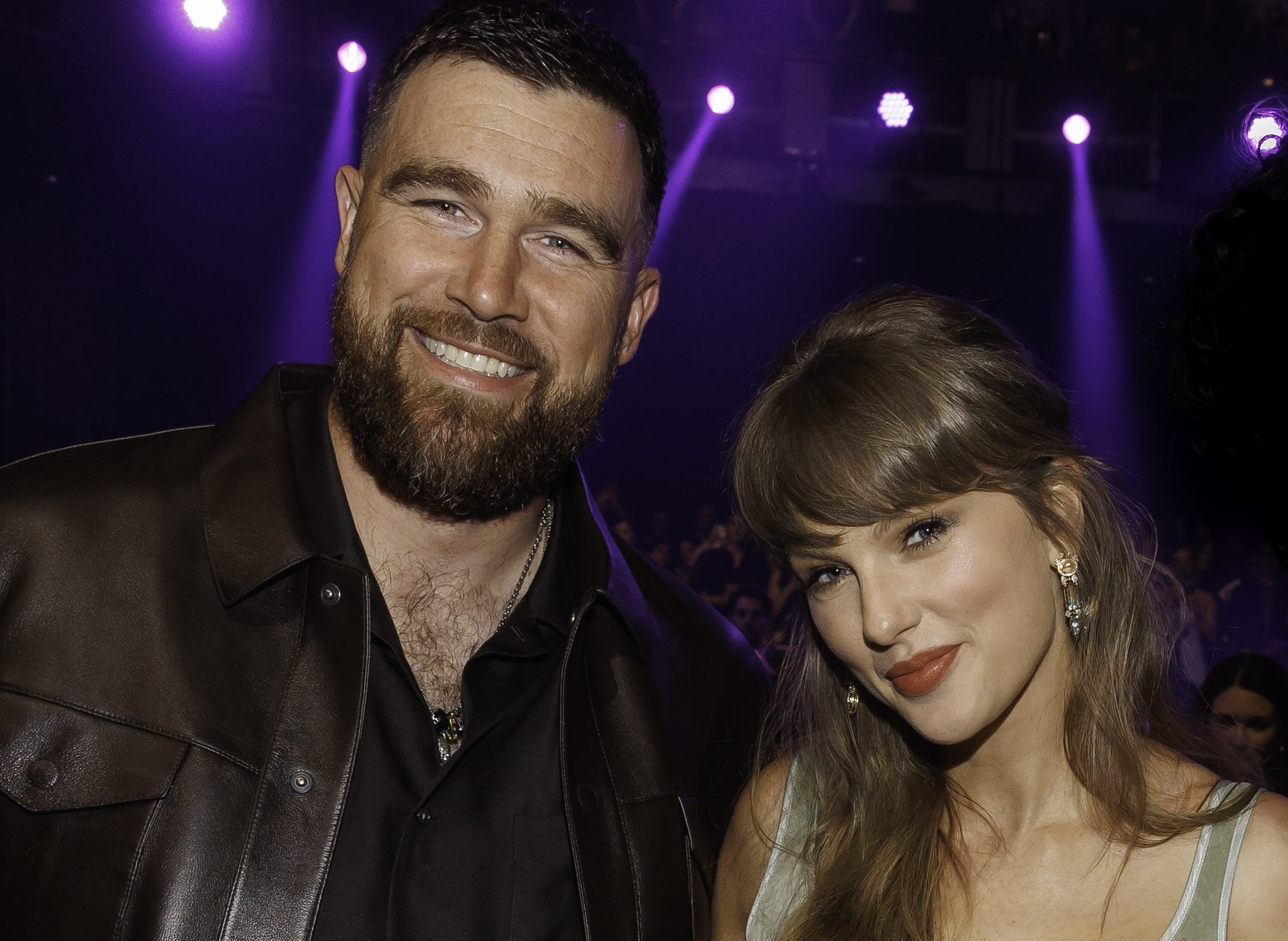 A bearded man in a black leather jacket and a woman with brunette hair and red lipstick pose together under purple stage lighting at an indoor event.