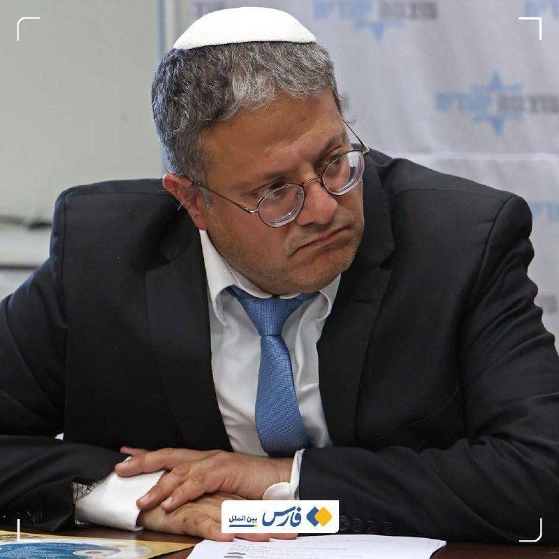 A man wearing glasses, a dark suit, blue tie, and a white kippah sits at a table with hands folded, looking downward beside documents.