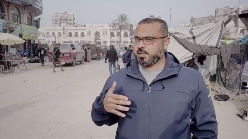 A bespectacled, bearded man in a blue jacket gestures with his hand while standing on a street with pedestrians, vehicles, tents, and buildings in the background.