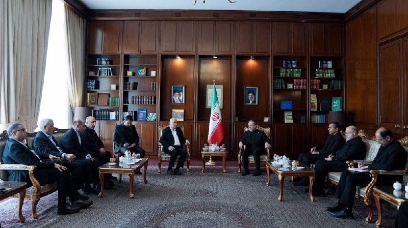 Group of men in suits seated in a wood-paneled room with bookshelves, an Iranian flag, and framed portraits.