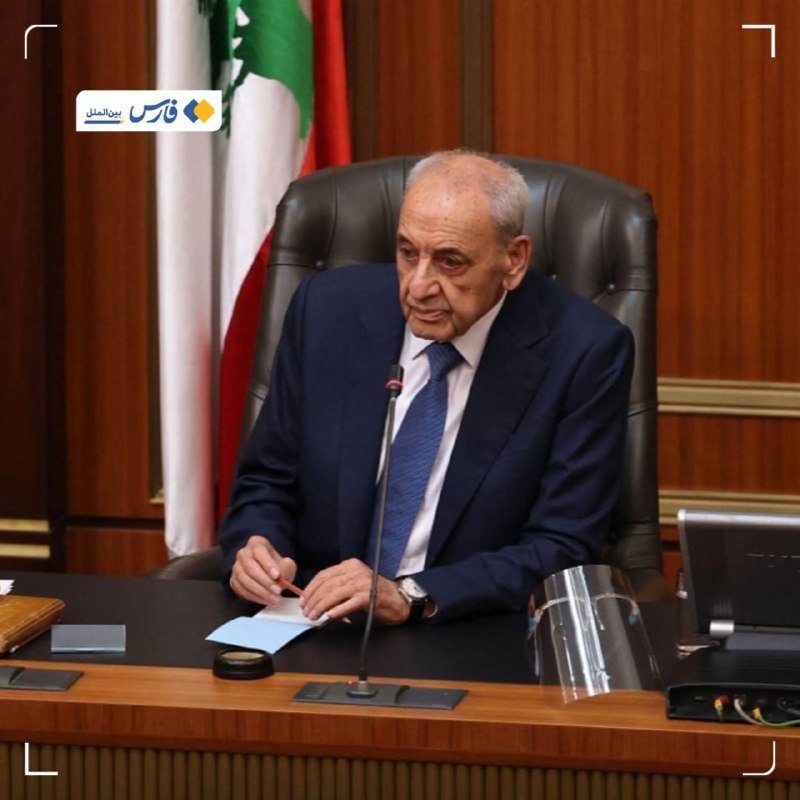 An older man in a dark suit and blue tie sits at a wooden desk with a microphone, in front of a Lebanese flag.