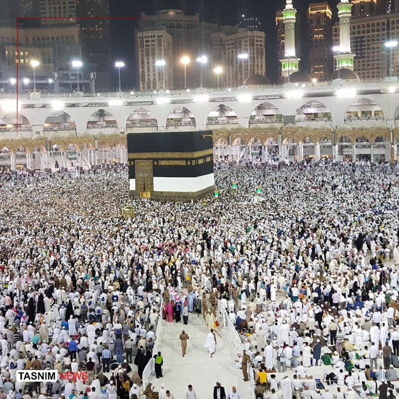 Thousands of people in white garments gather around a large black cubic structure at night, surrounded by illuminated arched architecture and minarets.