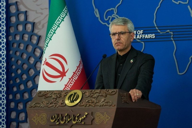 A man in a dark suit stands at a podium bearing the emblem of Iran, with the Iranian flag and a world map backdrop partially reading "Y OF…AFFAIRS."
