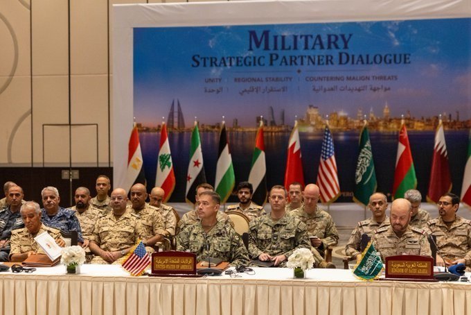 Military officials in camouflage uniforms sit at a table beneath a banner reading "Military Strategic Partner Dialogue," with multiple national flags displayed behind them.