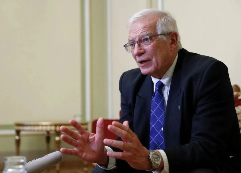 An older man with white hair and glasses, wearing a dark suit and blue patterned tie, gestures with both hands while speaking at a table with a microphone.